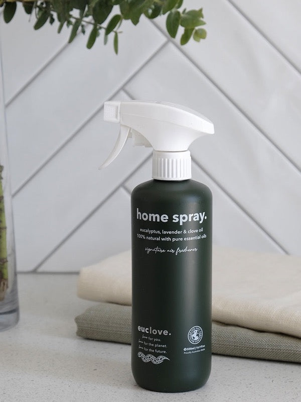 Dark green Euclove Home Spray bottle with a white trigger sprayer on a pale counter beside folded towels, with diagonal white tiles and green foliage behind.