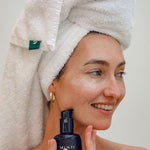Smiling woman in a towel turban holding the Mukti Hydrating Cleansing Lotion bottle to her cheek