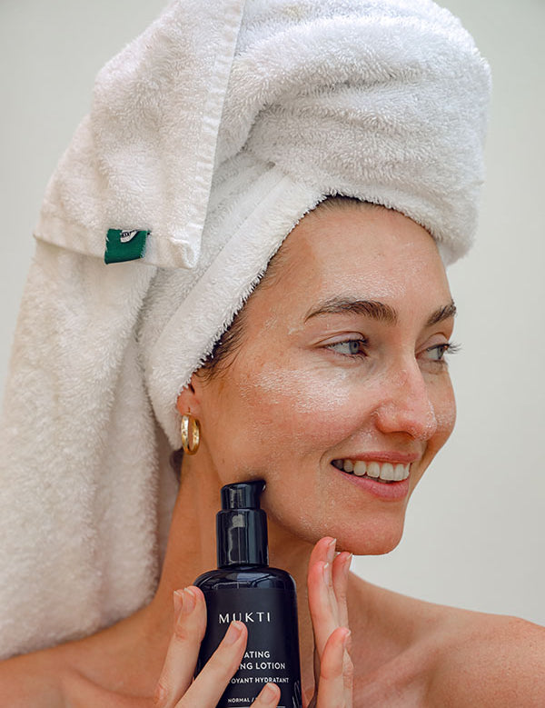 Smiling woman in a towel turban holding the Mukti Hydrating Cleansing Lotion bottle to her cheek