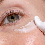 Close-up of a freckled eye area as the curved white applicator of INIKA Eye Cream spreads a stripe of white cream beneath the lower eyelid.