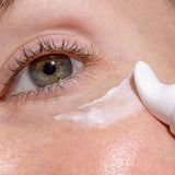 Close-up of a freckled eye area as the curved white applicator of INIKA Eye Cream spreads a stripe of white cream beneath the lower eyelid.