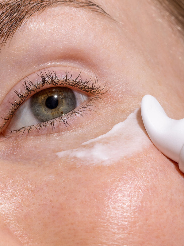 Close-up of a freckled eye area as the curved white applicator of INIKA Eye Cream spreads a stripe of white cream beneath the lower eyelid.
