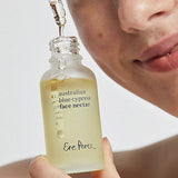 Woman holding an open bottle of Ere Perez Australian Blue Cypress Face Nectar as oil drips from the dropper