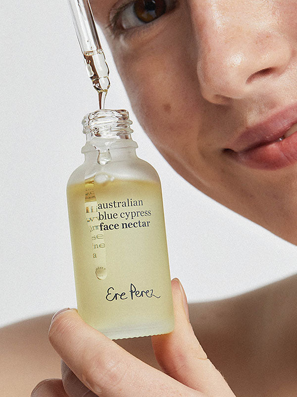 Woman holding an open bottle of Ere Perez Australian Blue Cypress Face Nectar as oil drips from the dropper