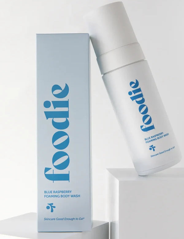 White Foodie Blue Raspberry Foaming Body Wash bottle leaning on a white display block beside its pale blue box, both with large blue lettering.