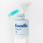 Close-up of Foodie Blueberry Burst Glow Serum 20ml on a white pedestal, with turquoise serum dripping from a dropper above the open bottle.