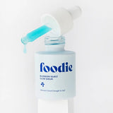 Close-up of Foodie Blueberry Burst Glow Serum 20ml on a white pedestal, with turquoise serum dripping from a dropper above the open bottle.
