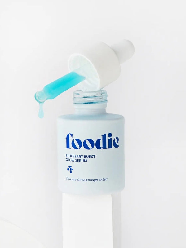 Close-up of Foodie Blueberry Burst Glow Serum 20ml on a white pedestal, with turquoise serum dripping from a dropper above the open bottle.