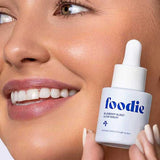 Smiling woman holding Foodie Blueberry Burst Glow Serum 20ml beside her glowing complexion.
