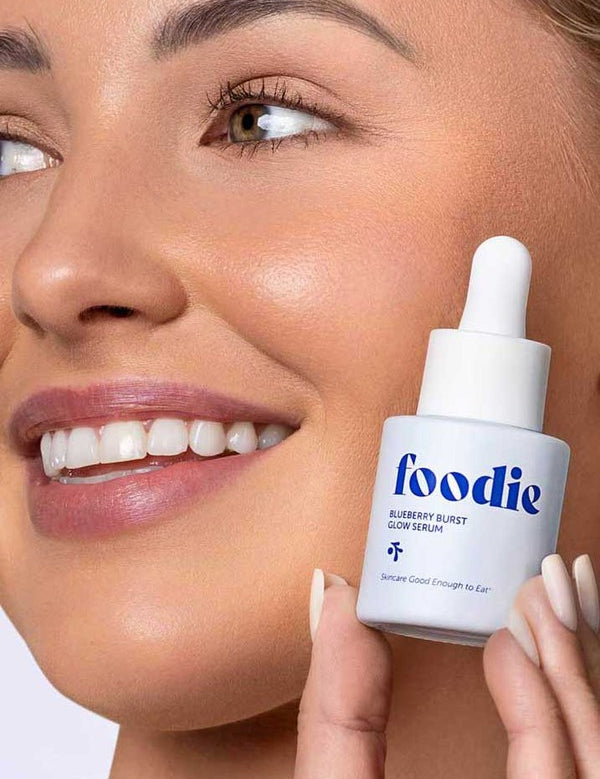 Smiling woman holding Foodie Blueberry Burst Glow Serum 20ml beside her glowing complexion.