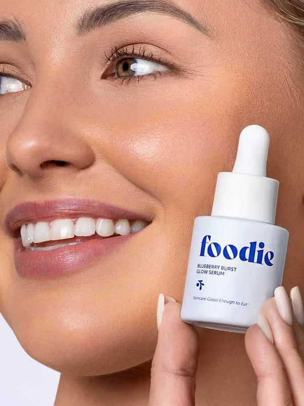 Smiling woman holding Foodie Blueberry Burst Glow Serum 20ml beside her glowing complexion.