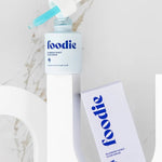 Foodie Blueberry Burst Glow Serum 20ml open bottle on a curved white display beside its lilac carton, with turquoise serum visible in the dropper against a marble background.