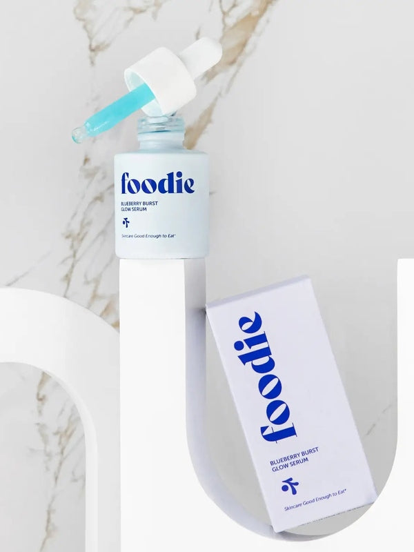 Foodie Blueberry Burst Glow Serum 20ml open bottle on a curved white display beside its lilac carton, with turquoise serum visible in the dropper against a marble background.