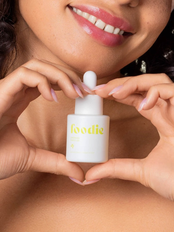 Vanessa Megan Foodie Lemon Aid Pimple Gel 20ml held beneath a smiling model's chin, with her fingers forming a heart around the yellow-labelled dropper bottle.