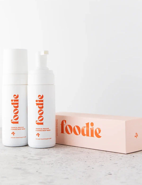Two white Foodie Tropical Pavlova Foaming Body Wash bottles beside a peach box on a pale stone surface; one bottle is capped and the other's pump is exposed.