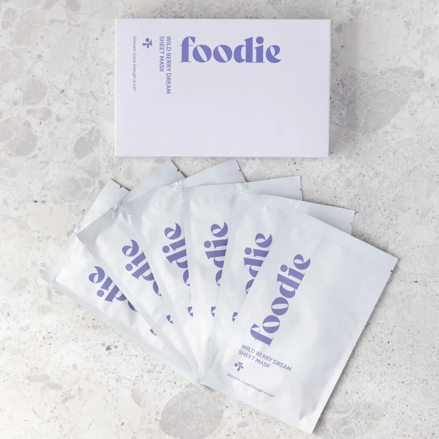 Overhead view of the Foodie Wild Berry Dream Sheet Mask 6-Pack, showing the lavender box above six overlapping white sachets with purple Foodie lettering on pale stone.