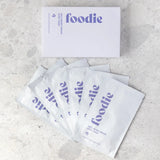 Overhead view of the Foodie Wild Berry Dream Sheet Mask 6-Pack, showing the lavender box above six overlapping white sachets with purple Foodie lettering on pale stone.