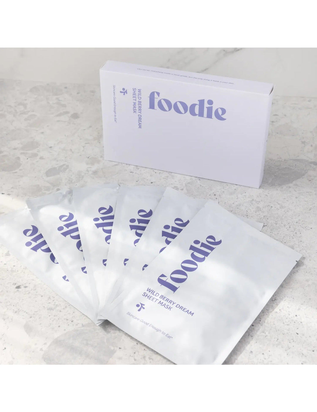 Foodie Wild Berry Dream Sheet Mask 6-Pack, with six white sachets fanned in front of the pale lavender box on a light stone surface, viewed at an angle.