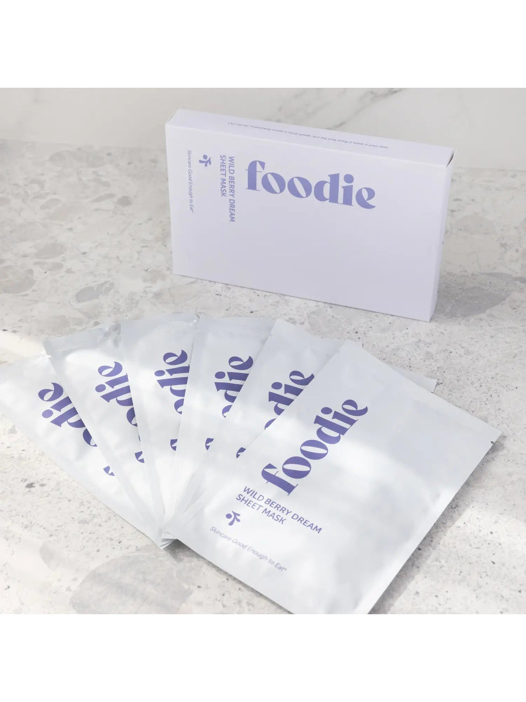 Foodie Wild Berry Dream Sheet Mask 6-Pack, with six white sachets fanned in front of the pale lavender box on a light stone surface, viewed at an angle.