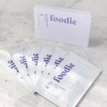 Foodie Wild Berry Dream Sheet Mask 6-Pack, with six white sachets fanned in front of the pale lavender box on a light stone surface, viewed at an angle.