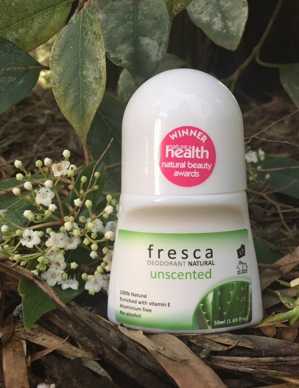 Fresca Unscented natural roll-on deodorant bottle standing among green leaves and small white flowers in an outdoor botanical setting.
