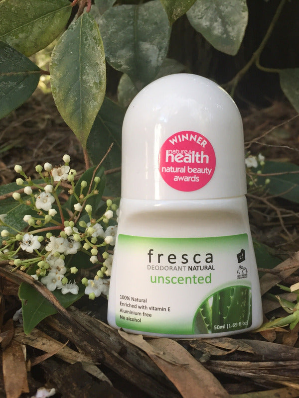 Fresca Unscented natural roll-on deodorant bottle standing among green leaves and small white flowers in an outdoor botanical setting.