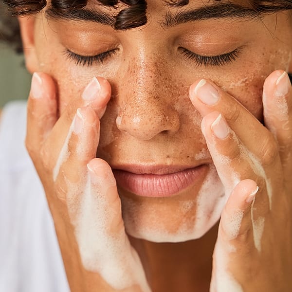 Dark skin woman with short curly hair with her eyes closed washing her face with a face cleansing foam.