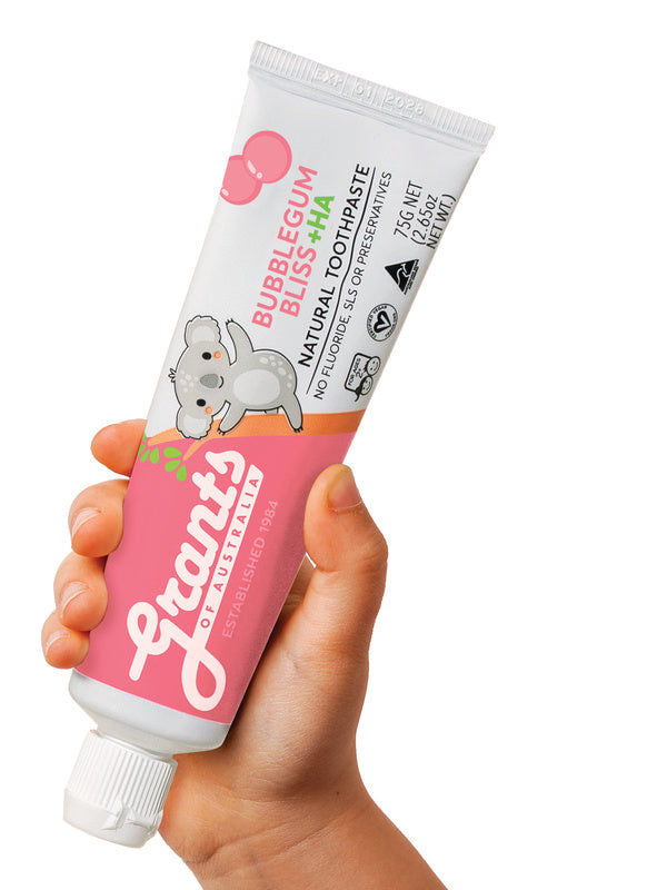 A hand holding a pink and white Grants Kids Bubblegum Bliss +HA 75g toothpaste tube with a koala illustration against a white background.