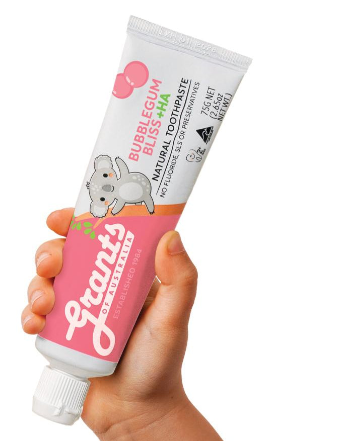 A hand holding a pink and white Grants Kids Bubblegum Bliss +HA 75g toothpaste tube with a koala illustration against a white background.