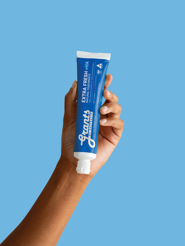 A hand holding a blue Grants Extra Fresh +HA 110g toothpaste tube against a blue background.