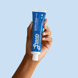 A hand holding a blue Grants Extra Fresh +HA 110g toothpaste tube against a pale blue background.