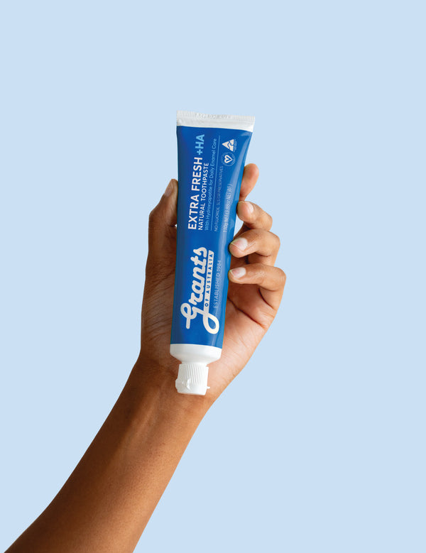 A hand holding a blue Grants Extra Fresh +HA 110g toothpaste tube against a pale blue background.