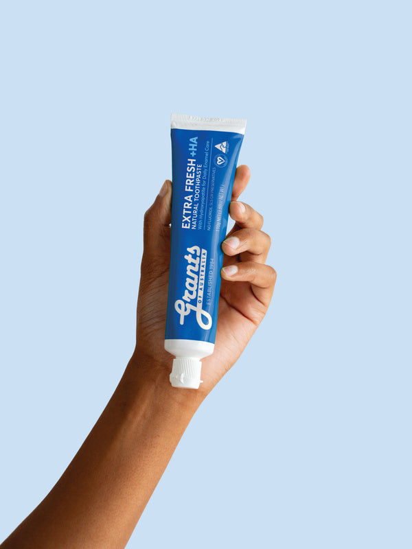 A hand holding a blue Grants Extra Fresh +HA 110g toothpaste tube against a pale blue background.