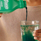 Hand pouring green Grants Liquid Chlorophyll into a clear glass of water held against a neutral knitted top.