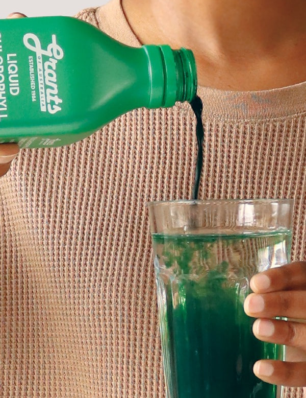 Hand pouring green Grants Liquid Chlorophyll into a clear glass of water held against a neutral knitted top.