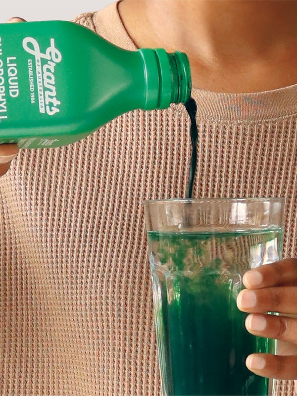 Hand pouring green Grants Liquid Chlorophyll into a clear glass of water held against a neutral knitted top.