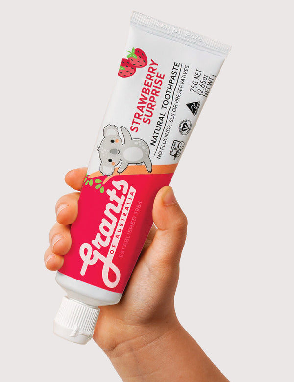 Hand holding a tube of Grants Kids Strawberry Surprise fluoride-free toothpaste against a pale background.