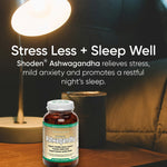 Green Nutritionals Shoden Ashwagandha bottle on a wooden bedside table beneath a glowing reading lamp, beside a dark mug and bed pillows. Overlaid text reads Stress Less + Sleep Well and promotes stress relief and restful sleep.