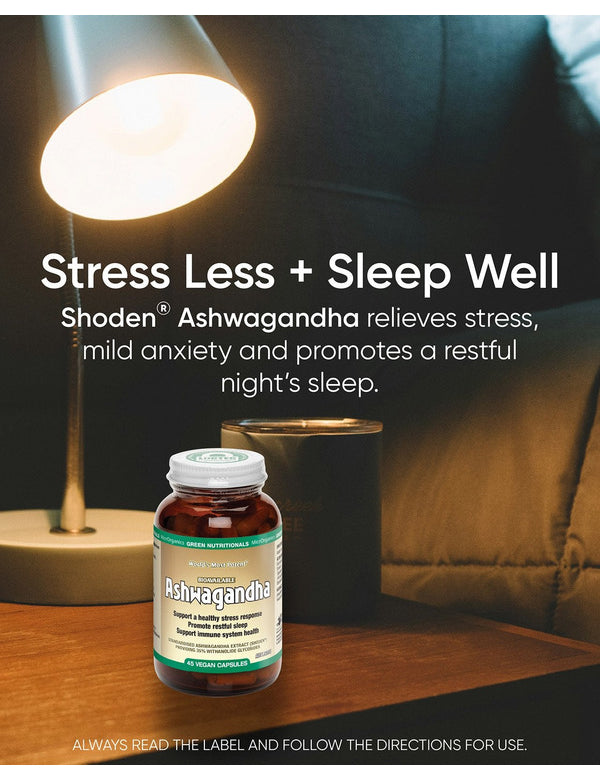 Green Nutritionals Shoden Ashwagandha bottle on a wooden bedside table beneath a glowing reading lamp, beside a dark mug and bed pillows. Overlaid text reads Stress Less + Sleep Well and promotes stress relief and restful sleep.