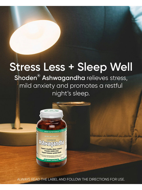 Green Nutritionals Shoden Ashwagandha bottle on a wooden bedside table beneath a glowing reading lamp, beside a dark mug and bed pillows. Overlaid text reads Stress Less + Sleep Well and promotes stress relief and restful sleep.