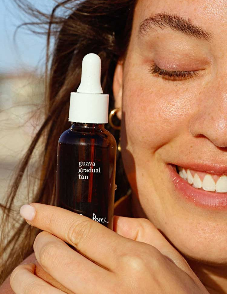 A smiling woman with glowing skin holds the Ere Perez Guava Gradual Tan dropper bottle near her face, basking in sunlight that highlights her clear complexion and natural sunless radiance.