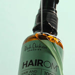 A thin stream of clear oil pours over a tilted amber HairOM bottle, running down its mint-green label beneath the black pump.