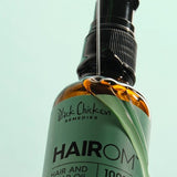 A thin stream of clear oil pours over a tilted amber HairOM bottle, running down its mint-green label beneath the black pump.