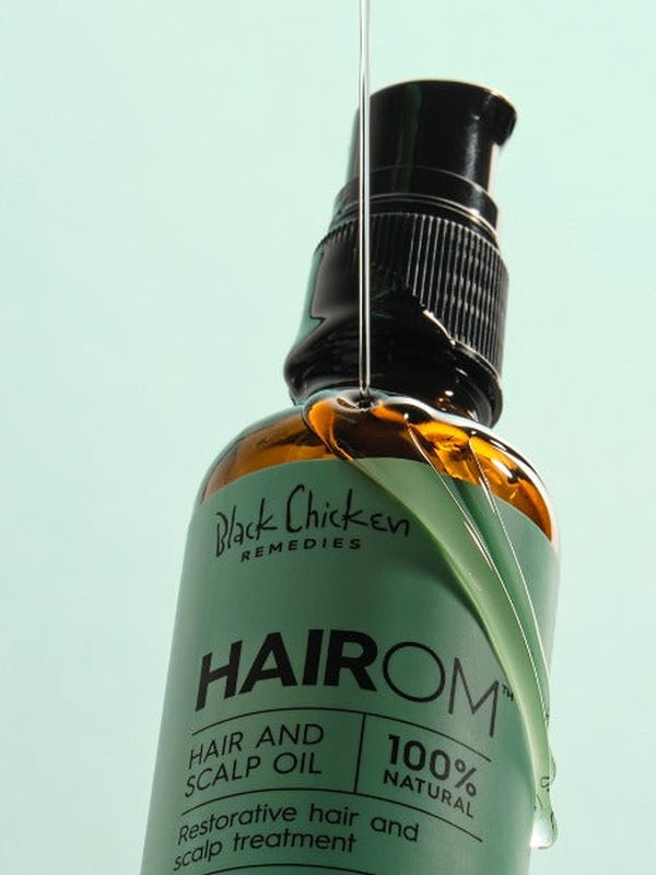 A thin stream of clear oil pours over a tilted amber HairOM bottle, running down its mint-green label beneath the black pump.