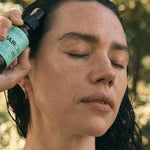 A woman with dark, wet hair closes her eyes and holds an amber HairOM pump bottle against her hairline outdoors.