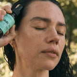 A woman with dark, wet hair closes her eyes and holds an amber HairOM pump bottle against her hairline outdoors.
