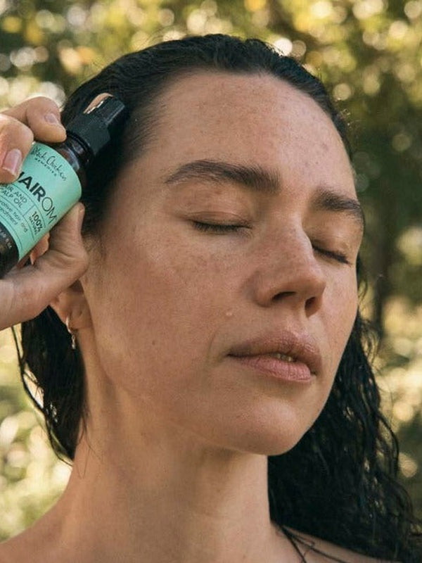 A woman with dark, wet hair closes her eyes and holds an amber HairOM pump bottle against her hairline outdoors.
