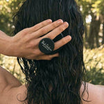 Rear view of a person using a round black scalp brush on long, dark, wet hair outdoors, with the brush handle held between their fingers.