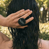 Rear view of a person using a round black scalp brush on long, dark, wet hair outdoors, with the brush handle held between their fingers.