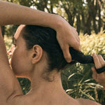 Rear view of a person gathering dark, wet hair at the nape of the neck with both hands, outdoors among green plants.